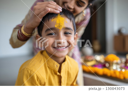 Boy with yellow powder on forehead smiling during traditional ceremony with woman blessing him indoors 130843073