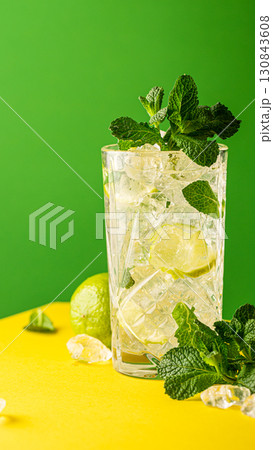 Refreshing cocktail with ice, mint leaves, and lime on vibrant yellow and green background 130843608