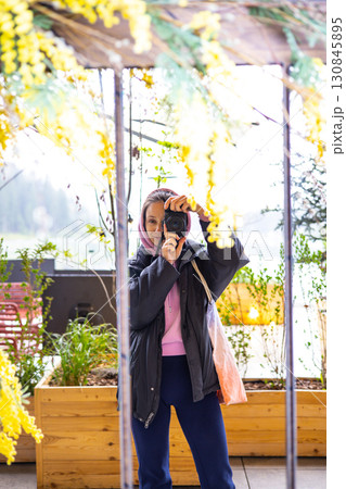 A woman capturing a moment with a camera in front of a mirror, surrounded by yellow mimosa flowers. A young girl takes a picture of herself reflection A woman capturing a moment with a camera in front of a mirror, surrounded by yellow mimosa flowers. A young girl takes a picture of herself reflection 130845895