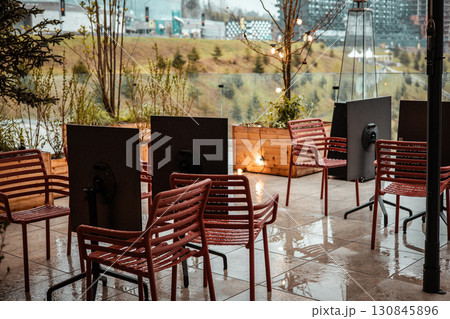 Empty patio chairs, tables sit in a rain-swept outdoor setting with a city view. An outdoor terrace captured in an overcast day. String lights garland Empty patio chairs, tables sit in a rain-swept outdoor setting with a city view. An outdoor terrace captured in an overcast day. String lights garland 130845896
