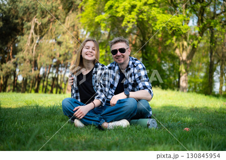 Happy Father And Daughter Sit On The Grass In The Park, Looking Into The Camera And Enjoying The Outdoors 130845954
