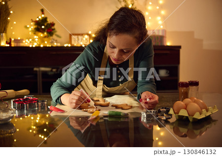 Woman Decorating Gingerbread Cookies in Festive Kitchen Setting With Christmas Lights 130846132