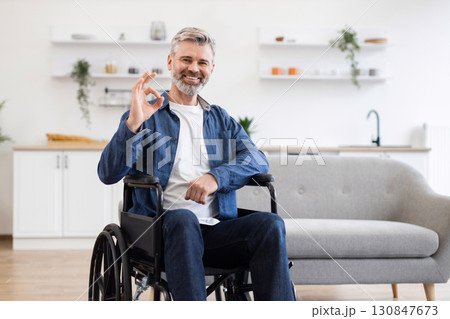 Senior Caucasian male in wheelchair smiling, making okay gesture, sitting in modern bright kitchen. Image depicts confidence, inclusivity, positivity, and accessibility in a home environment. 130847673