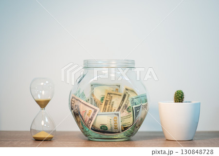 Glass jar with money on the table, sand clock and cactus nearby. Concept of savings, collecting money for something 130848728