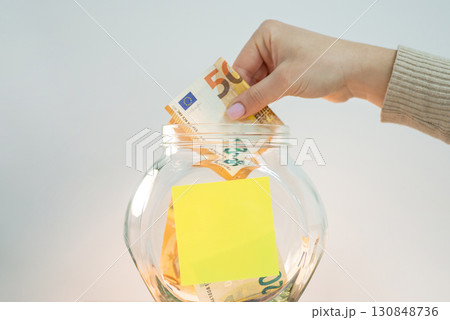 Female hand putting 50 euro banknote into glass jar, empty sticky note for inscription on it. Concept of savings Female hand putting 50 euro banknote into glass jar, empty sticky note for inscription on it. Concept of savings 130848736