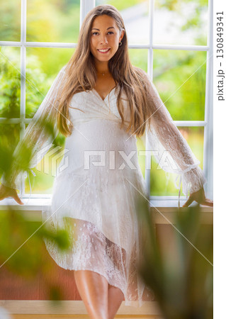 Pregnant woman smiling standing by a window in natural light Pregnant woman smiling standing by a window in natural light 130849341