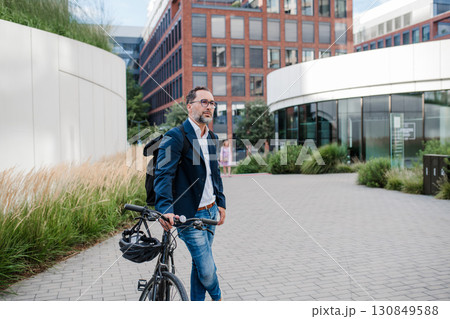 City commuter walking with bike to office, wearing backpack. 130849588