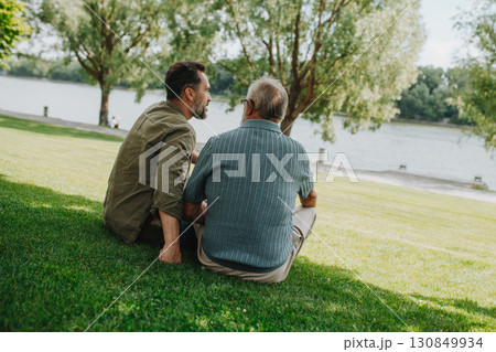 Father and adult son sitting on grass in park, sharing peaceful moment. 130849934
