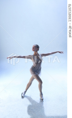 Graceful figure skater extending arms in balanced pose under soft light 130851070