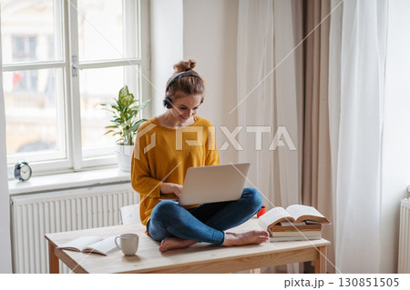 Young female student sitting at the table, using headphones while studying. 130851505