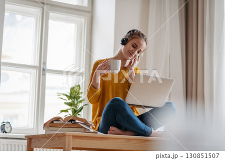 Young female student sitting at the table, using headphones while studying. 130851507