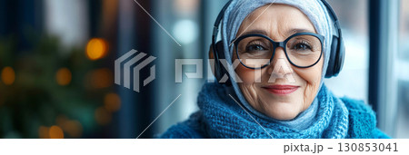 A relaxed, smiling Arab grandma with headphones stands in front of a navy blue background, embodying chic elegance in a simple, minimalist mock-up design, banner A relaxed, smiling Arab grandma with headphones stands in front of a navy blue background, embodying chic elegance in a simple, minimalist mock-up design, banner 130853041