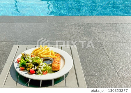 Summer vacation. Plate of grilled salmon, salad, and fries sits poolside, symbolizing relaxed healthy dining during sun-soaked summer vacation at upscale resort. Healthy lifestyle Summer vacation. Plate of grilled salmon, salad, and fries sits poolside, symbolizing relaxed healthy dining during sun-soaked summer vacation at upscale resort. Healthy lifestyle 130853277