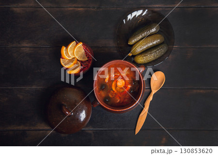 Homemade Traditional Russian soup Solyanka with meat, sausages, vegetables, pickles and olives with lemon in ceramic pot with wooden spoon on dark wooden background, 130853620