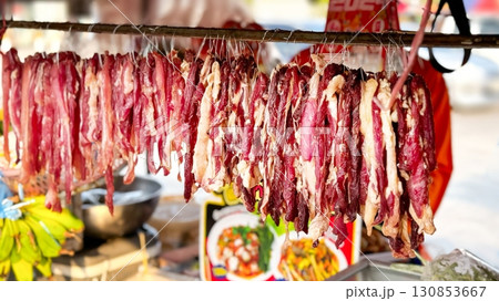 Dried meat hangs in an open-air market, a rustic Asian delicacy offered amidst a backdrop of fresh produce, inviting passersby to indulge in local culinary traditions. Street food Dried meat hangs in an open-air market, a rustic Asian delicacy offered amidst a backdrop of fresh produce, inviting passersby to indulge in local culinary traditions. Street food 130853667