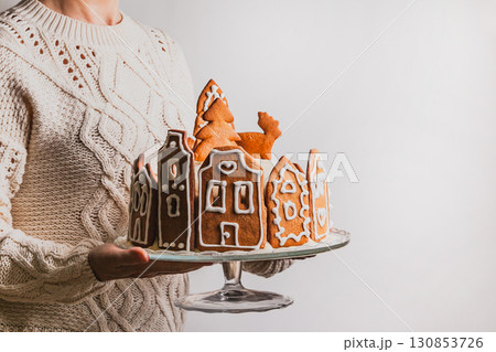 Woman holds homemade cake with gingerbread city house, Christmas tree and reindeer on glass stand Woman holds homemade cake with gingerbread city house, Christmas tree and reindeer on glass stand 130853726
