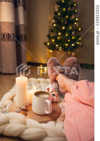 Women's feet in home fur slippers, cup of cappuccino, candles on blanket of thick yarn and Christmas tree with lights. 130853832