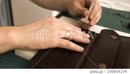 Punching stitching holes in brown leather wallet with pricking iron in artisan workshop 130854174