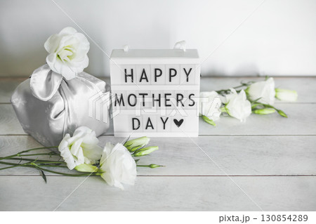 Light box with lettering Happy Mother's Day, Gift box trendy wrapped in silk fabric in Furoshiki technique, white flowers Eustoma or Lisianthus on light wooden background with copy space. 130854289