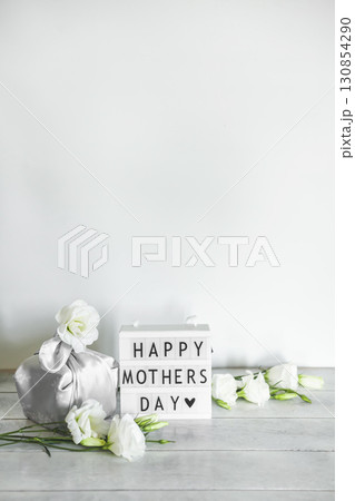 Light box with lettering Happy Mother's Day, Gift box trendy wrapped in silk fabric in Furoshiki technique, white flowers Eustoma or Lisianthus on light wooden background with copy space. 130854290