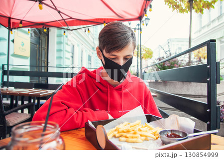 Wearing of masks in public places. Young man sits in a street cafe in a protective mask and looks at French fries Wearing of masks in public places. Young man sits in a street cafe in a protective mask and looks at French fries 130854999