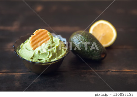 Traditional latinamerican mexican sauce guacamole in bowl with cracker, avocado and lemon on dark background Traditional latinamerican mexican sauce guacamole in bowl with cracker, avocado and lemon on dark background 130855155