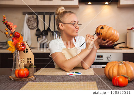 Woman on kitchen paints pumpkin for Halloween in room with autumn decor and lamp house. Cozy home and preparation for Halloween Woman on kitchen paints pumpkin for Halloween in room with autumn decor and lamp house. Cozy home and preparation for Halloween 130855511