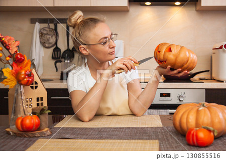 Woman on kitchen carves a pumpkin for Halloween in a room with autumn decor and a lamp house. Cosy home and preparing for Halloween Woman on kitchen carves a pumpkin for Halloween in a room with autumn decor and a lamp house. Cosy home and preparing for Halloween 130855516