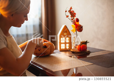 Woman carves pumpkin for Halloween in room with autumn decor and lamp house. Cosy home and preparing for Halloween. 130855521