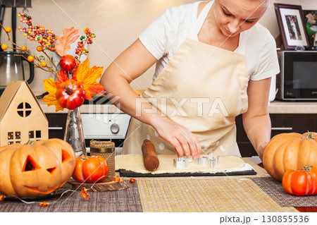 A woman cuts out cookies for Halloween with a mold in the kitchen with an autumn decor. A woman cuts out cookies for Halloween with a mold in the kitchen with an autumn decor. 130855536