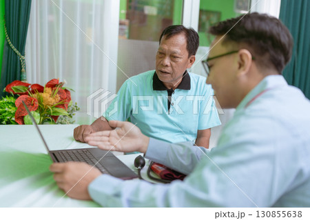 In a bright room, a doctor engages in consultation with a patient using a laptop In a bright room, a doctor engages in consultation with a patient using a laptop 130855638