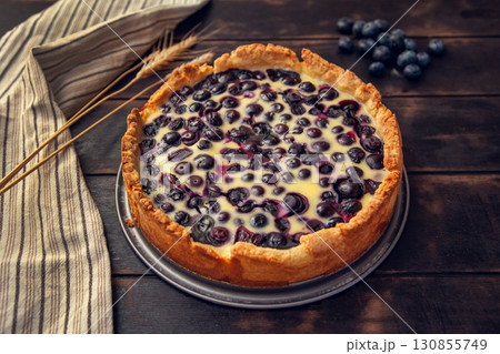 Rustic shortbread pie with blueberries in sour cream filling on a wooden background with kitchen towel and wheat ears 130855749