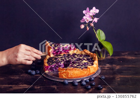 Womans hand cuts off and takes a piece of rustic shortbread pie with blueberries in sour cream filling on a wooden background and phalaenopsis flower. Close up Womans hand cuts off and takes a piece of rustic shortbread pie with blueberries in sour cream filling on a wooden background and phalaenopsis flower. Close up 130855762
