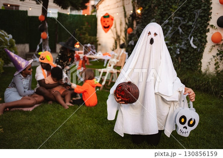 Child wearing ghost costume holding pumpkin and skull trick or treat bag during Halloween party in the garden Child wearing ghost costume holding pumpkin and skull trick or treat bag during Halloween party in the garden 130856159