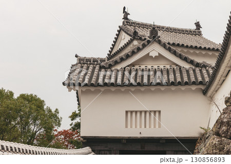 Architectural detail of Himeji Castle with tiled roofs stone walls and foliage under seasonal sky 130856893