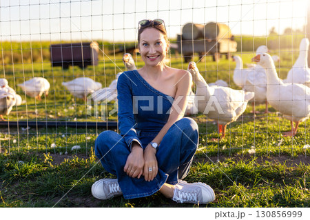 Portrait young adult happy smiling woman wear jeans denim jumpsuit enjoy having fun countryside farm animal geese flock warm sunset light. Female person rural country village farmyard bird poultry Portrait young adult happy smiling woman wear jeans denim jumpsuit enjoy having fun countryside farm animal geese flock warm sunset light. Female person rural country village farmyard bird poultry 130856999