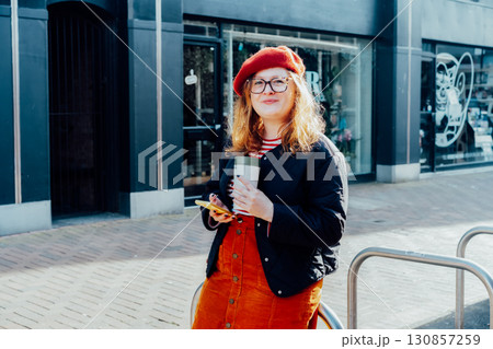 Young smiling plus size woman with backpack using mobile phone, drinking coffee outdoors on the street. Confident fashion hipster messaging, chatting, working remotely. Using navigator app for travel. 130857259
