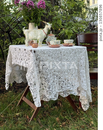 Lace tablecloth draped over table with teapot and cups in garden setting Lace tablecloth draped over table with teapot and cups in garden setting 130857896