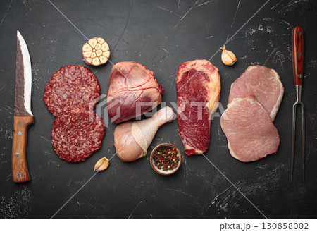 Selection of assorted raw meat cuts for zero carb carnivore diet: uncooked beef steak, pork meat, ground meat patty, heart, and chicken legs on black stone background top view 130858002