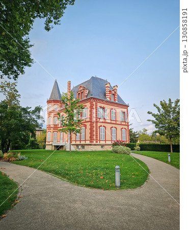 The Castel Rose in L'Isle-Adam, France. Historic mansion characterized by its pink walls and distinctive red brick trim. The building is surrounded by a lush green lawn and winding paths 130858191