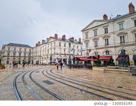Place Sainte-Croix in Orleans, France. Grand, wet cobblestone square during a rainy day. Ornate buildings and outdoor cafes. Tram tracks curve through the foreground, and people with umbrellas walking 130858193