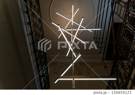 A close-up of a contemporary light fixture featuring multiple intersecting light tubes suspended in a stairwell. 130859125