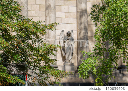 A sculpture of an angel with large wings, carved from stone, is located on the facade of an ancient building between two columns. The figure seems to embrace itself. It is partially hidden by green tr A sculpture of an angel with large wings, carved from stone, is located on the facade of an ancient building between two columns. The figure seems to embrace itself. It is partially hidden by green tr 130859608