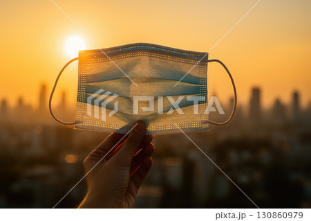 Surgical mask held by hand against bright sunset with city skyline background, symbolizing health care and pandemic safety awareness in urban environment 130860979
