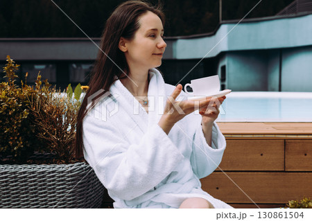 A relaxed woman enjoys cup of tea near a pool, wearing a white robe in a peaceful setting. A serene girl enjoys a moment with a cup of morning coffee. A relaxed woman enjoys cup of tea near a pool, wearing a white robe in a peaceful setting. A serene girl enjoys a moment with a cup of morning coffee. 130861504