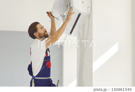 Professional electrician man with screwdriver maintaining modern air conditioner indoors. Professional electrician man with screwdriver maintaining modern air conditioner indoors. 130861540