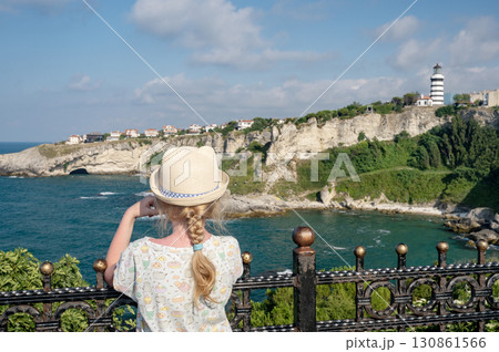 A girl in a hat near a decorative fence looks at a rock with a lighthouse over the sea A girl in a hat near a decorative fence looks at a rock with a lighthouse over the sea 130861566
