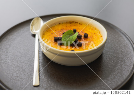 A creamy creme brulee, topped with chocolate and a fresh mint leaf. A delicious dessert on a dark plate with a silver spoon, ready to be savored. A creamy creme brulee, topped with chocolate and a fresh mint leaf. A delicious dessert on a dark plate with a silver spoon, ready to be savored. 130862141