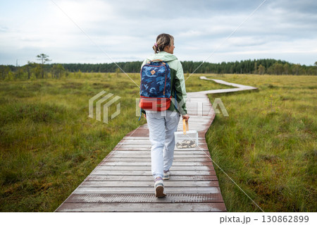 Rearview woman forager moving along marked eco trail in wetland woodland carrying harvest basket 130862899