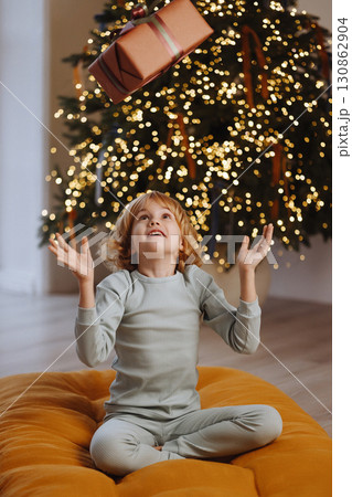 Smiling girl in pajamas tossing gift box near Christmas tree with lights in cozy room 130862904